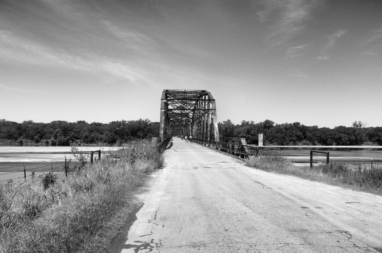 B&W cyclists crossing an old bridge.